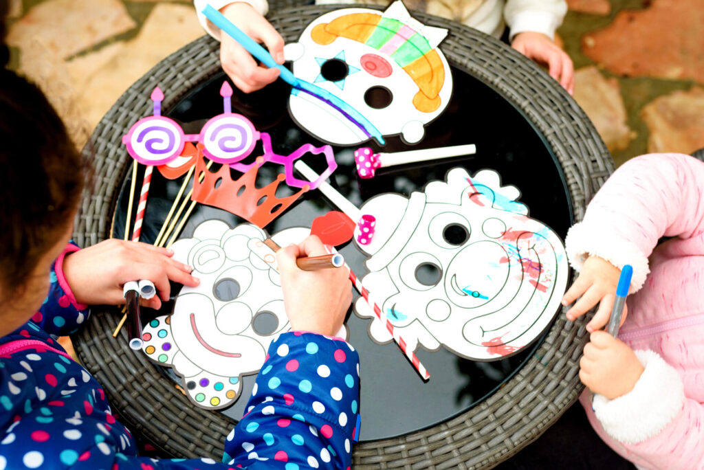 Kinderen knutselen met stiften carnavalsmaskers aan een ronde tafel, terwijl feestelijke accessoires zoals brillen en kronen op tafel liggen