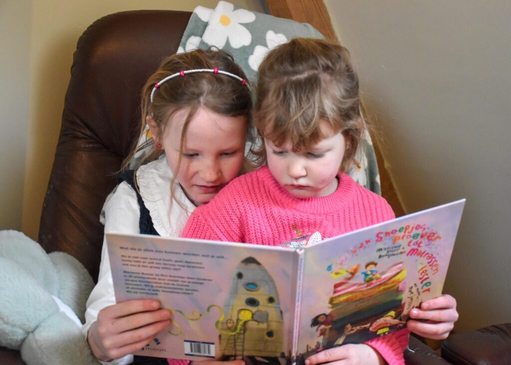 Twee zussen zitten samen knus in een stoel en lezen aandachtig het boek Van Snoepjesproever tot Matrassentester.