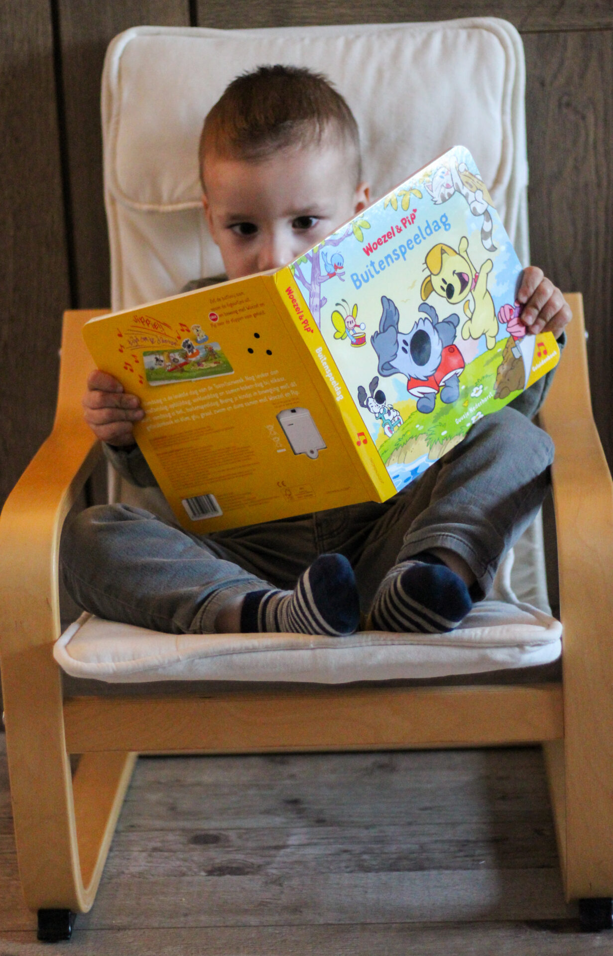 Jongen zit met opgetrokken benen in een stoel en bladert geconcentreerd door Woezel & Pip: Buitenspeeldag.