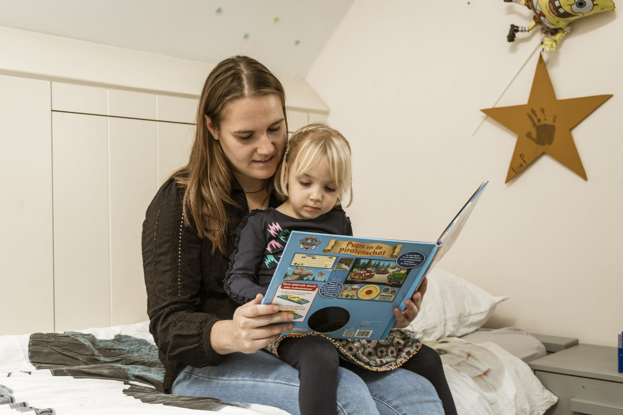 Moeder leest op bed voor aan haar peuterdochter met een kleurrijk kinderboek over pups en piraten.