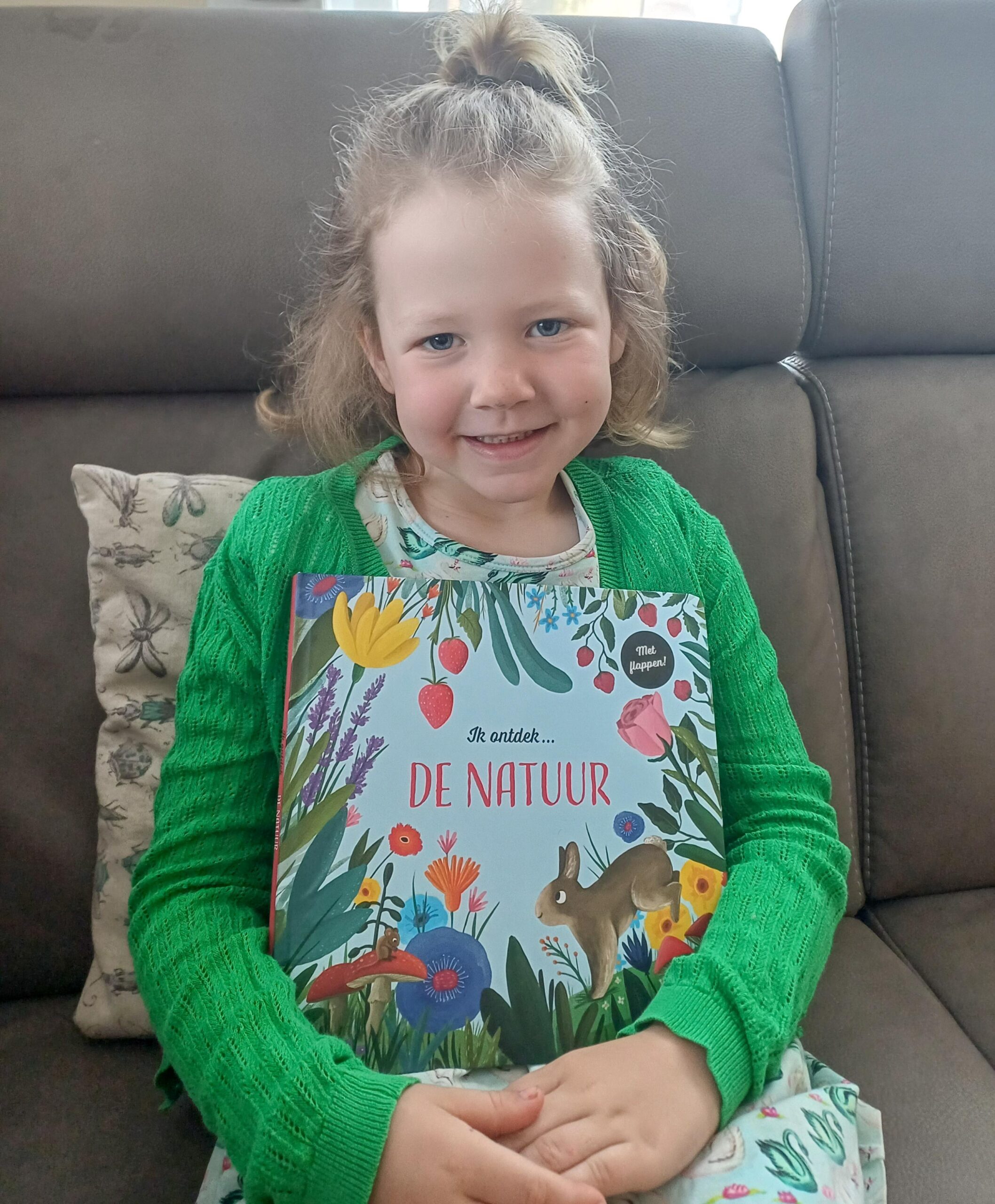 Lachend meisje met groen vest houdt trots het boek “Ik ontdek de natuur” op schoot terwijl ze op een bank zit.