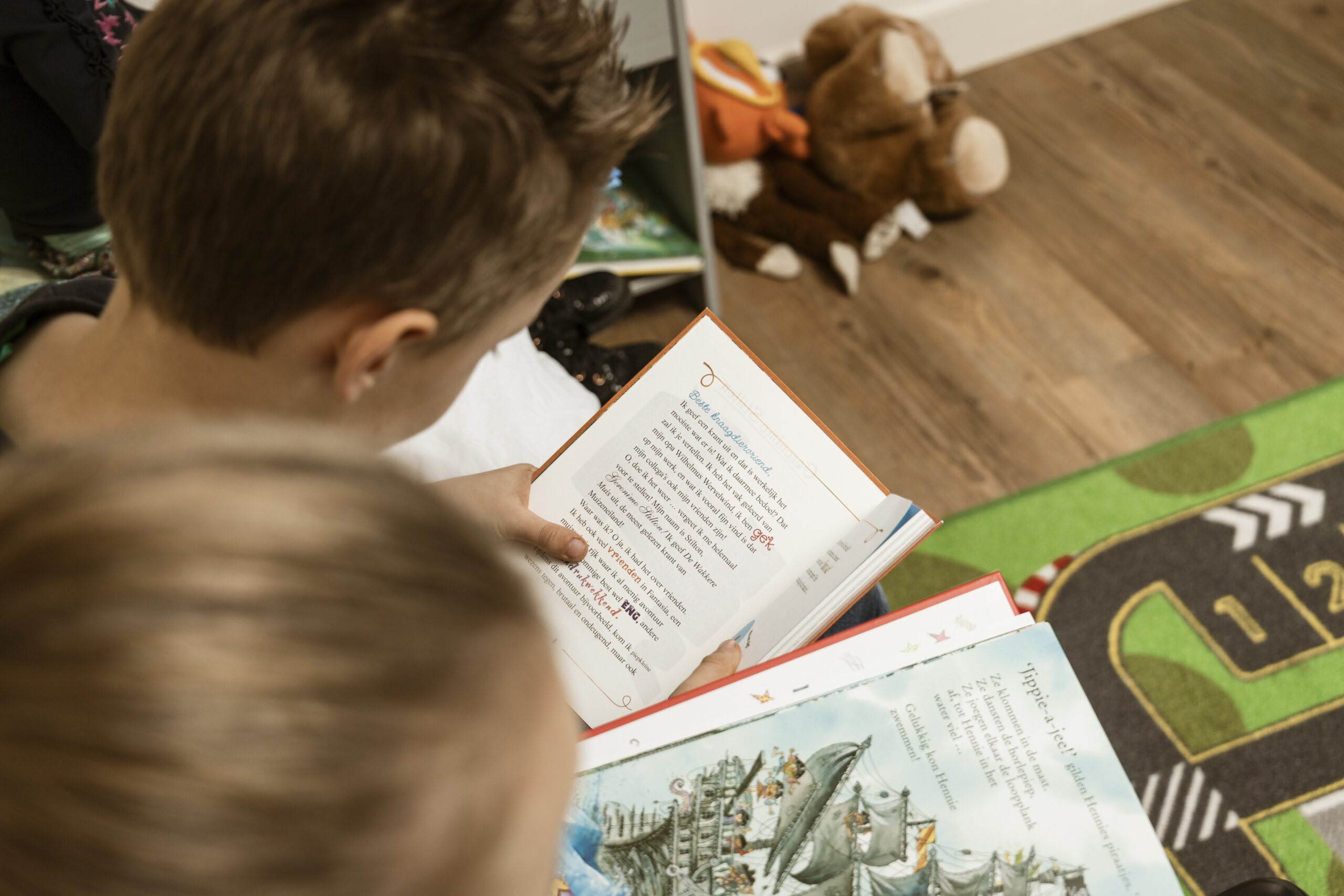 Twee kinderen lezen samen een kinderboek op de grond, met knuffels en een speelkleed op de achtergrond.