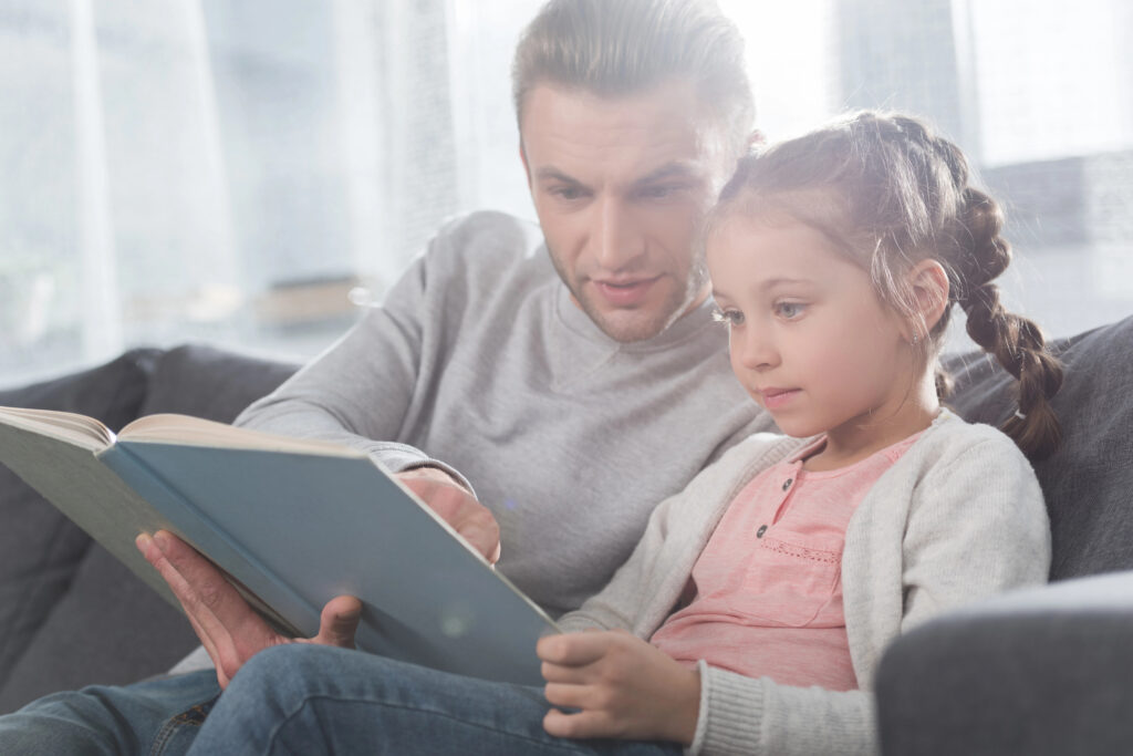 Vader leest samen met dochter een educatief kinderboekje thuis op de bank